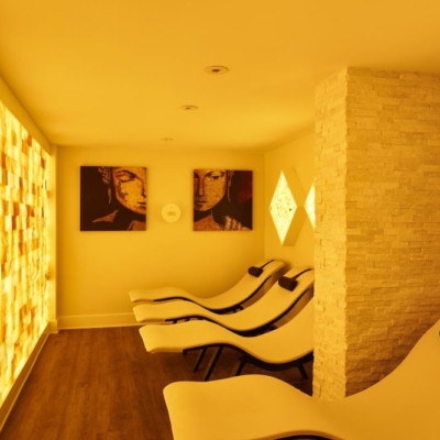 Three White Chaises With Brown Headrests Facing A Orange Backlit Salt Stone Wall With Three Diamond Shaped Salt Stone Décor And Two Paintings With Faces.