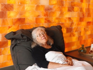 Woman In A Black Shirt Relaxing Under A White Blanket In Front Of A Himalayan Salt Panel Wall.