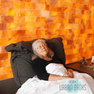 Woman In A Black Shirt Relaxing On A Black Chair With A White Blanket Covering Her In A Front Of A Himalayan Salt Stone Wall With The Text In The Lower Right Saying &Quot;Asha Urban Baths&Quot;.
