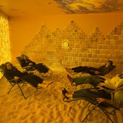 Five Women Smiling In Reclined Chairs On A Salt Covered Floor With A Led Backlit Salt Panel And Square Salt Brick Walls At  Prana Salt Cave - Wilmington, Nc.