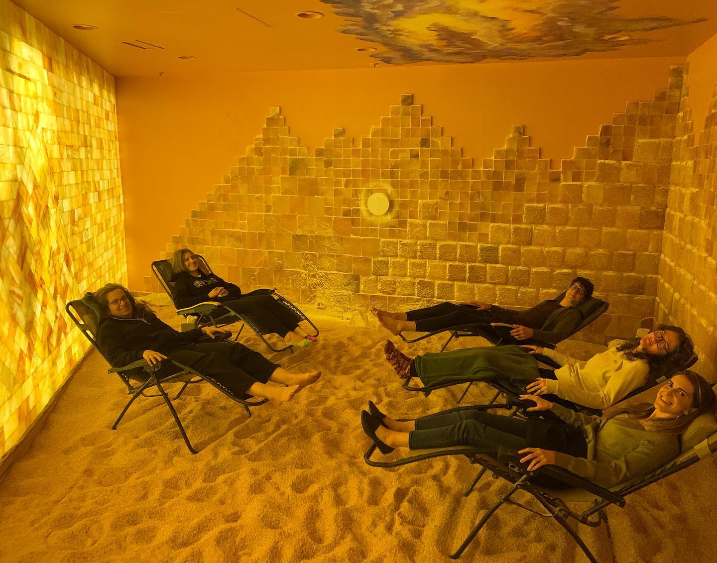 Five Women Smiling In Reclined Chairs On A Salt Covered Floor With A Led Backlit Salt Panel And Square Salt Brick Walls At  Prana Salt Cave - Wilmington, Nc.