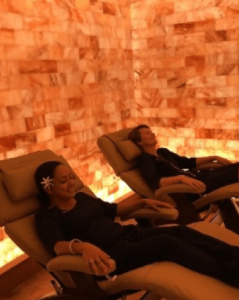 A Man And A Woman Laying On Recliners In A Salt Room With Himalayan Salt Brick Walls For Décor.