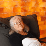 A Senior Woman Sitting In A Chair During A Salt Therapy Session With A Himalayan Salt Brick Wall.