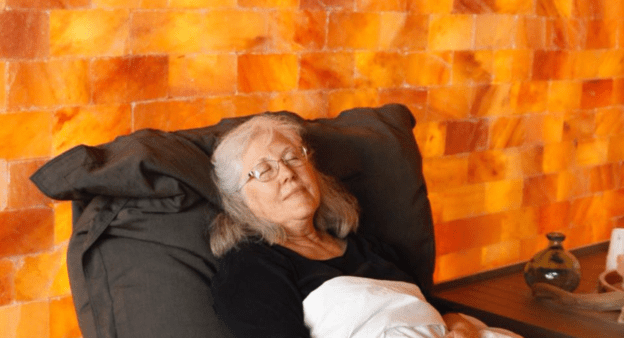A senior woman sitting in a chair during a salt therapy session with a Himalayan salt brick wall.