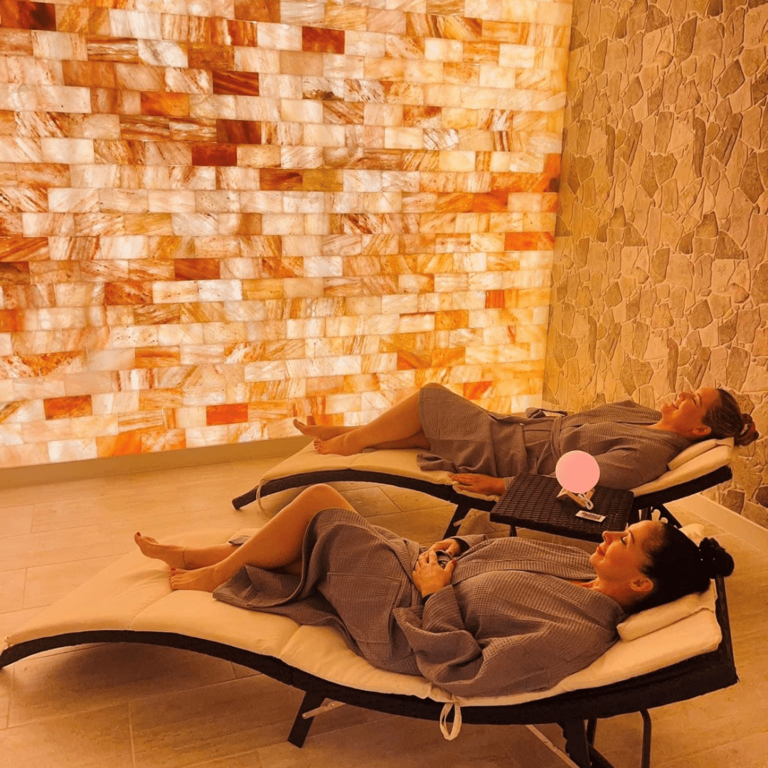 A salt room with two woman lying on reclining chairs with a Himalayan salt brick wall.